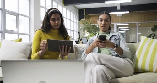 Diverse women colleagues comparing tablet charts, printed graphs at office lounge after phone alert