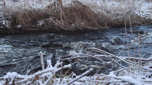 Nahaufnahmen von dunklem Flusswasser, das im Winter schnell fließt, mit Schnee an grasbewachsenen Ufern.