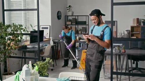 Male Cleaner Checking Phone while Female Colleague Mopping Floor in Office