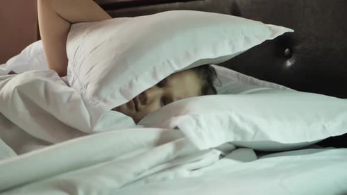 A Child Girl Uses White Pillows in Her Bed to Cover Her Ears From the Loud Noise That Disturbs the