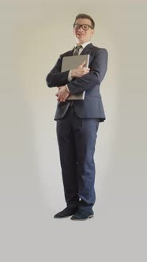 An Adult Man in a Business Suit and Glasses Stands and Holds a Laptop in His Hands on a White