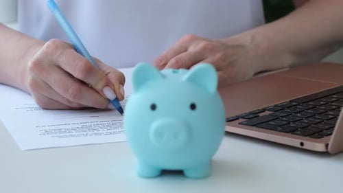 Woman Signs Document to Open Account or Deposit Next to Laptop and Piggy Bank