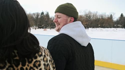Girl and guy in warm winter are ice-skating holding hands laughing enjoying date in park.