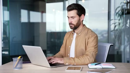 Focused handsome businessman typing on a laptop sitting at a workplace in a business office. Bearded