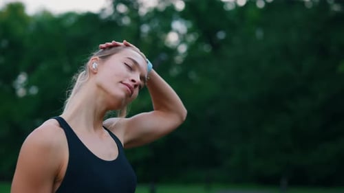 Sporty Caucasian Woman Stretch Her Neck Wearing Earphones Standing in the Park