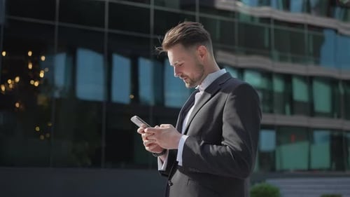 Young Businessman Holding Phone Reading Email Stands on Street Smiling Writes Message Communicates