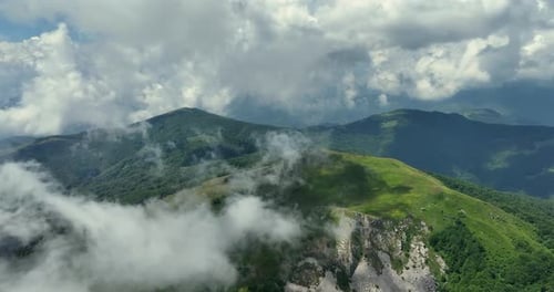 Flying trough white fluffy clouds above green mountain peaks. Beautiful summer sunny day on the high