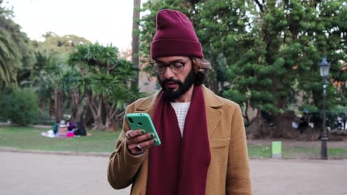 Happy Bearded Male Hipster Typing Messages with a Mobile Phone Social Media App at City Park