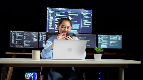 Programadora asiática usando smartphone enquanto escreve código em um laptop usando vários monitores