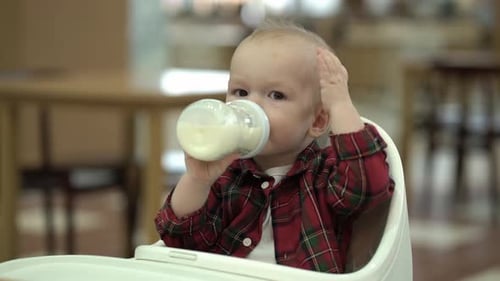 Baby Drinks Milk from a Bottle in High Chair