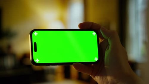 Hand Holding Modern Smartphone with Bright Green Chroma Key Screen in Dimly Lit Indoor Environment