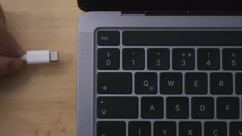 A top-down close-up of a person plug in a white USB-C cable to the laptop. The video focuses on