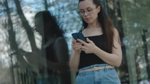 Young Woman with Eyeglasses Standing Alone in Front of a Building Using Mobile Phone Looking at