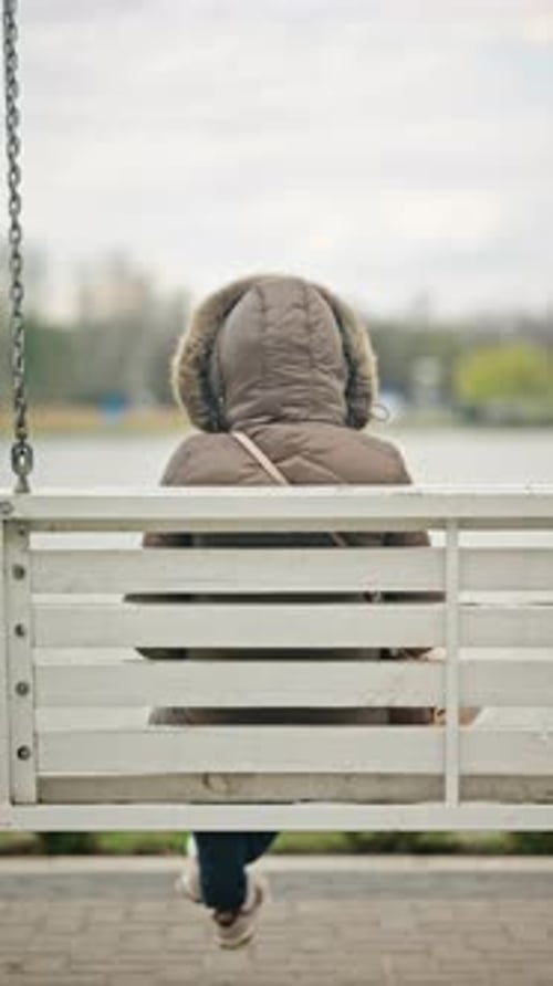 Back view of a woman sitting alone on a white wooden swing bench suspended by chains, facing a lake