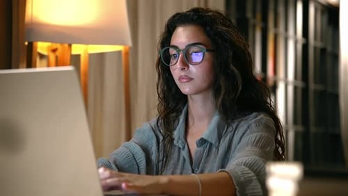 Young Woman in Home Office Looks at Laptop Screen and Works Typing on Keyboard