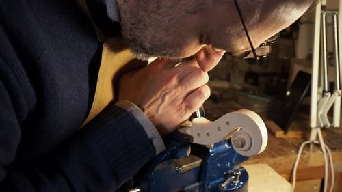Focused artisan wearing glasses leans close to the violin scroll held in a vise, carefully carving