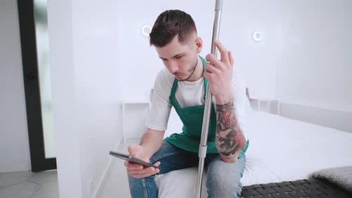 Young Cleaner in Green Apron Sitting on Bed Checking His Phone and Holding Mop for Cleaning in