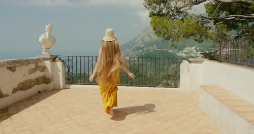 A woman runs on a terrace, looking out at the coastal landscape, with a marble bust nearby.