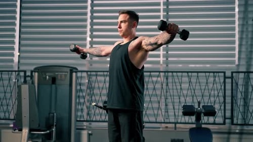 in the Gym Young Handsome Guy with Tattoos in a Black Tshirt Does Exercises with Two Dumbbells