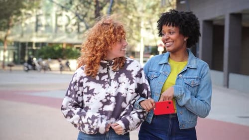 Two Cheerful Women Walk and Chat on City Sidewalk