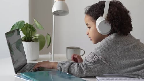 Primary School African American Student Child with Headphones Hand Writing in Book Using Laptop