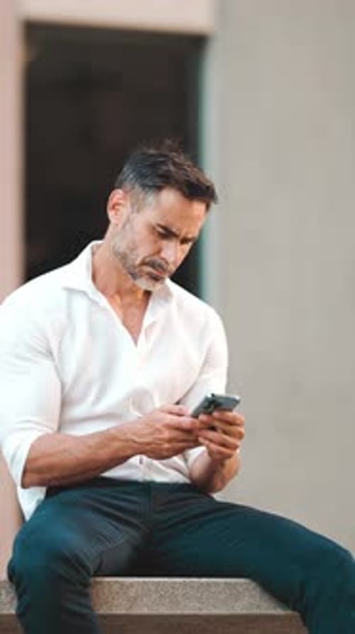 Mature businessman uses mobile phone sits on bench in the financial district in the city