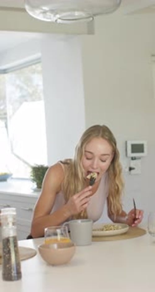 Vertical video: Adult woman at counter placing plate mug, cutting with knife, using fork, eating