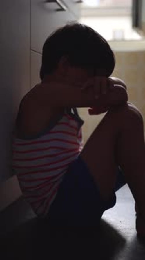 Child sitting on kitchen floor with head resting on arms expressing deeper emotional distress
