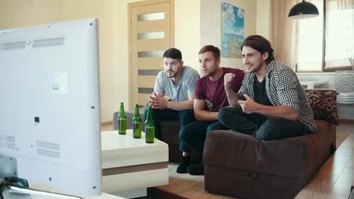 Three adult male friends with beer on table watching soccer indoors and celebrating victory