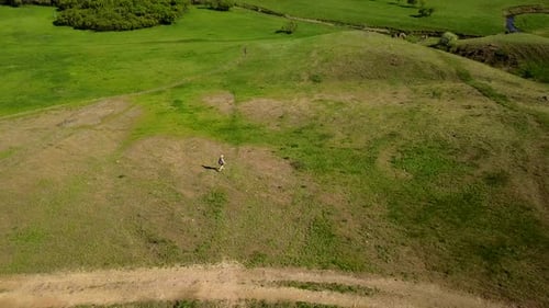 Aerial Video of Man Walking in Picturesque Valley on Green Grass Towards a River Drone Tracking Shot