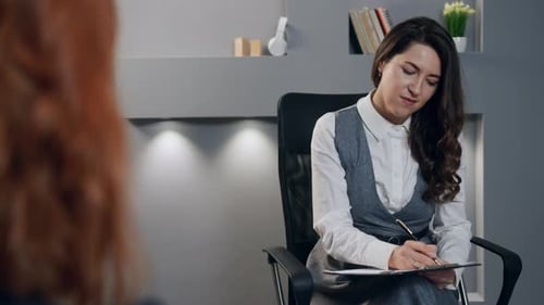 The Young Woman Psychologist Woman is Sitting on Chair and Notes Information From Red Hair Patient
