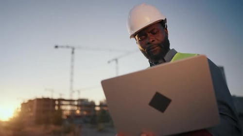 Schwarzer Mann mit weißem Helm auf dem Kopf hält modernes Laptopporträt eines Vorarbeiters auf der Baustelle