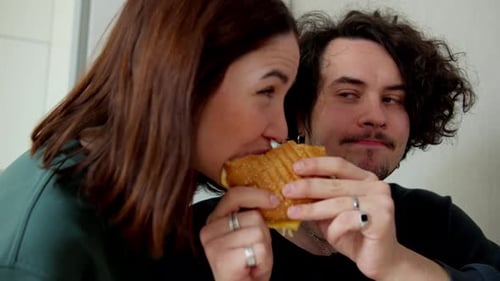 A Brunette Girl in a Green Jacket Bites a Sandwich That Her Boyfriend is Holding in His Hands Who