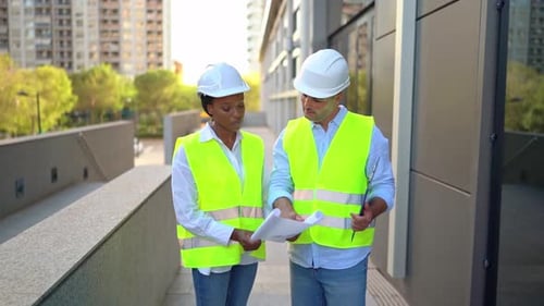 Two diverse engineers in hard hats and vests outside office examining building site with blueprint