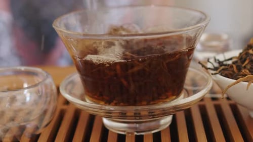 Panning shot of tea brewing in glass gaiwan standing on a wooden tray. Boiling water flowing into tr