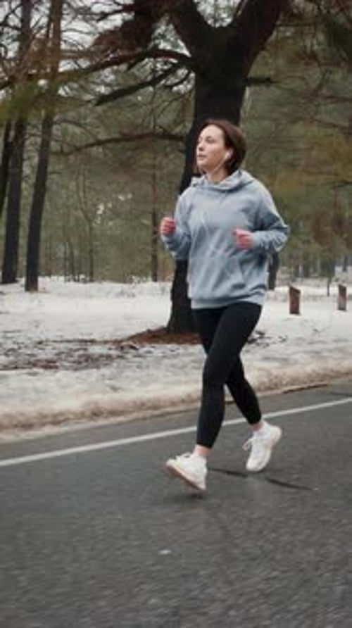 Fit female in sport clothes and earphones running in snowy park. Vertical video