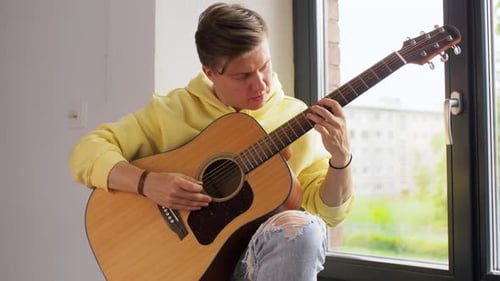 Young man tuning guitar by the window at home enjoying music and leisure