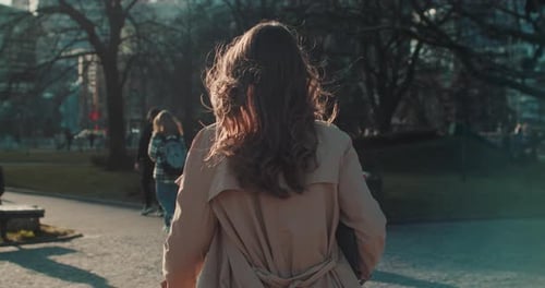 Camera Follows Beautiful Woman in Beige Coat with Long Brown Hair Walking Alone City Park Looking