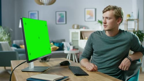 Handsome Young Man Works on a Green Mock-up Screen Personal Computer while Sitting at His Desk in t