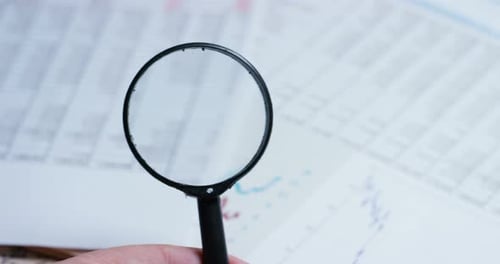 A hand with a magnifying glass passes over a financial newspaper and financial bag with a business