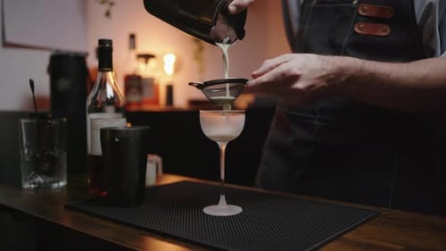 Skilled bartender crafting a cocktail at home bar. Pouring cocktail through strainer from shaker int