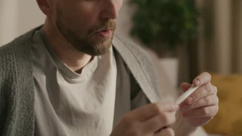 Man Looks at Thermometer, Coughing in Home Interior
