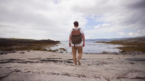 A Lone Traveler with a Backpack is Seen Walking Along a Sandy Beach in the Mountainous Region