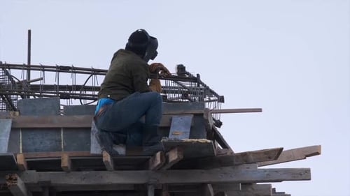 Man with helmet welds metal structure at height with flying sparks against sky background