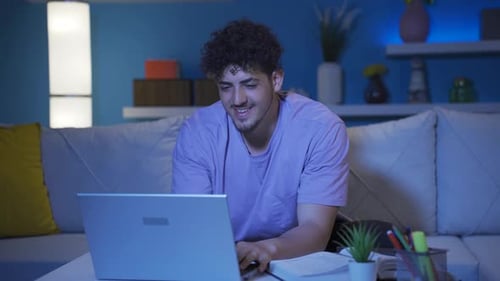 Young college student looking at laptop at night and smiling.