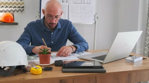 Architect Making Notes on Building Plans at Office Desk