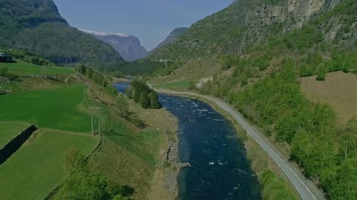 Aerial backwards shot of blue river surrounded by greened mountains and snowy peak in background - S