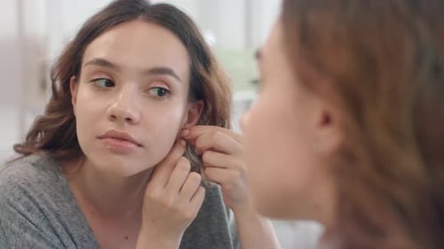Smiling Woman Puts on Earrings in Front of Mirror