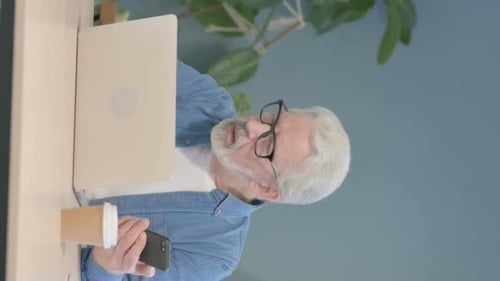 Senior Man Uses Smartphone and Laptop in Office
