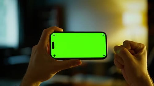 Person Holding Smartphone with Bright Green Chroma Key Display in Dimly Lit Indoor Room Blank Screen